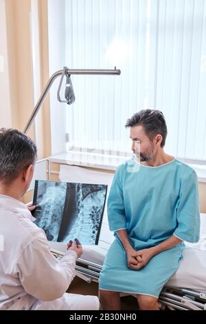 Sad mature patient with cancer sitting on bed and listening to doctors recommendations while doctor viewing x-ray image Stock Photo