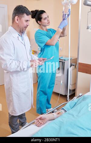 Young nurse preparing IV bag while giving patient IV, mature doctor talking to patient while viewing his test results on tablet Stock Photo