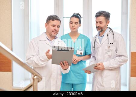 Mature doctor in lab coat holding folder and showing test results to colleagues while discussing symptoms of illness Stock Photo