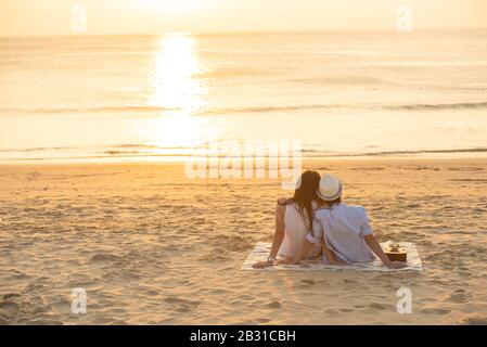 Couple in love watching sunset together on beach travel summer holidays. People silhouette from behind sitting enjoying view sunset sea on tropical Stock Photo