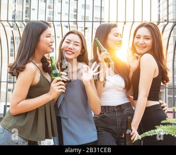 Group of young beautiful happy asian women holding bottle of beer chat together with friends while celebrating dance party on outdoor rooftop nightclu Stock Photo