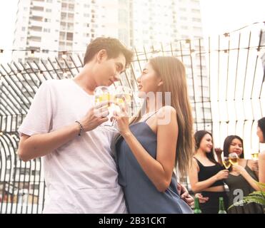 Young asian couple lover have fun dancing and drinking in night party on rooftop floor nightclub hand holding beer bottle and eye contact flirting at