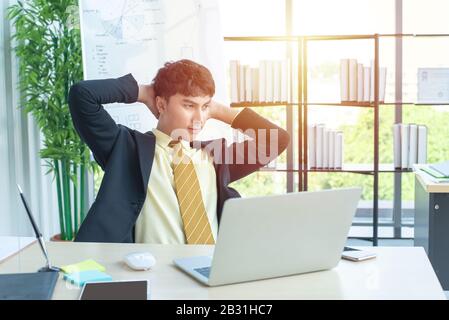 happy young asian businessman in formal dress sitting on chair with relax in modern office with computer laptop on table in workplace. Stock Photo