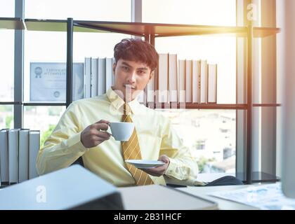 Happy young asian businessman in formal drink coffee sitting in modern workplace in break time Stock Photo