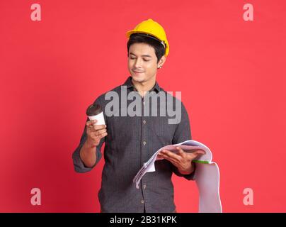 Young handsome asian man in casual dress with yellow hardhat hand holding paperwork blueprint and paper coffee cup  in architect and engineer worker Stock Photo