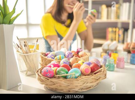 Focused on basket of decorated easter eggs with blurred of beautiful young asian woman painting easter eggs on table with decorated eggs for celebrate Stock Photo