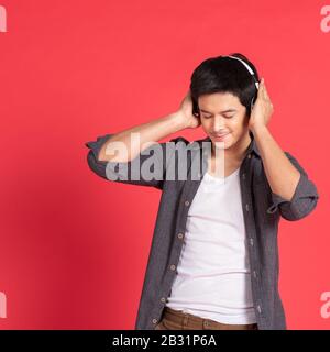 Image of happy young asian student man dress in casual shirt singing and listening to music with cell phone and wireless earphones isolated over red w Stock Photo