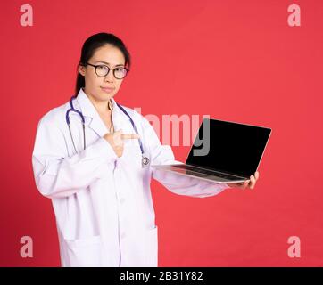 Portrait of smiling beautiful asian young woman doctor with digital computer laptop isolated on red background Stock Photo