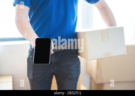 Young delivery man in uniform holding and showing smartphone with blank screen.Ai transportation technology concept. Stock Photo