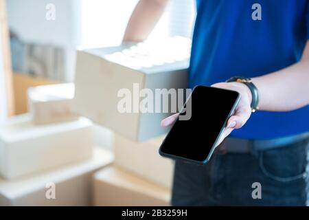 Young delivery man in uniform holding and showing smartphone with blank screen.Ai transportation technology concept. Stock Photo