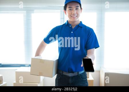 Young delivery man in uniform holding and showing smartphone with blank screen.Ai transportation technology concept. Stock Photo