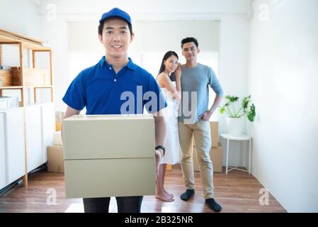Smiling young asian delivery man in blue uniform holding and carrying two cardbox parcel look at camera with happy young asia couple sender standing i Stock Photo