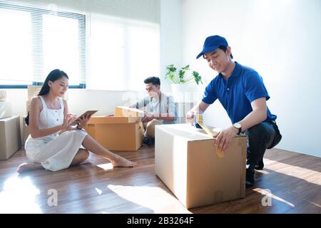 Delivery service worker man in blue uniform help happy couple customer packing parcel with tape in customer home.Woman with tablet signing proof docum Stock Photo