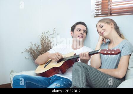 Happy young handsome caucasian man plays guitar and sing-song for his girlfriend sit on the sofa in the living room at the apartment. Stock Photo