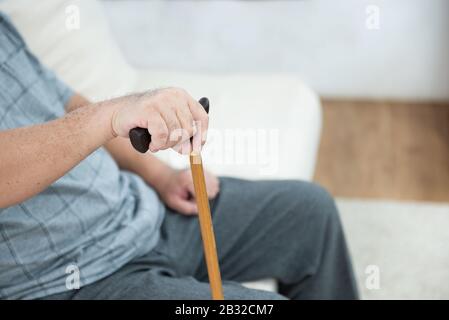 elderly old senior man sitting, resting his hands on wooden walking stick sitting on couch in living room in house after retirement. Stock Photo