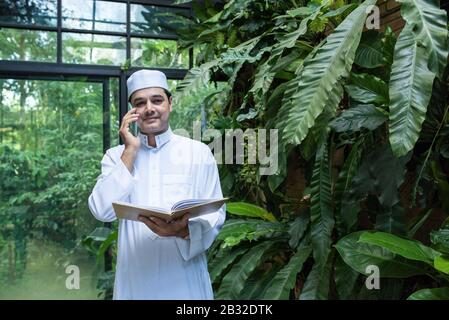 Middle eastern Emirati man in traditional clothing called kandura sitting on chair in coffee shop and talking with his mobile hand holding book.Smart Stock Photo