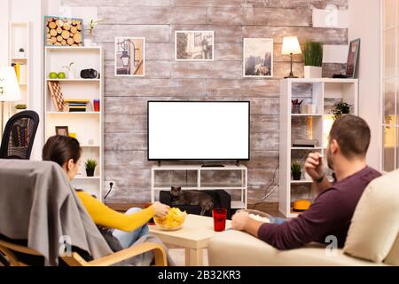 Couple looking at isolated TV screen in cozy living room while eating takeaway food Stock Photo