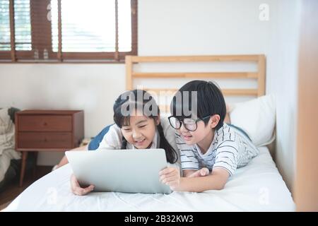 Happy asian boy and girl playing game in laptop lying on bed in bedroom in house,brother and sister using notebook do homework,education concept. Stock Photo