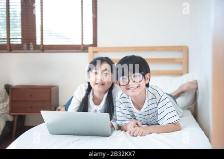Happy asian boy and girl playing game in laptop lying on bed in bedroom in house,brother and sister using notebook do homework,education concept. Stock Photo