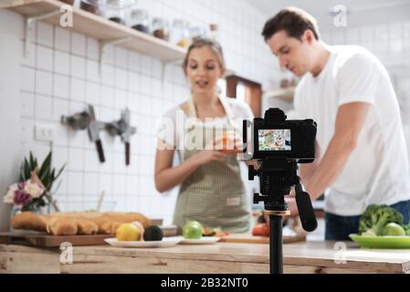 Professional digital mirrorless camera  recording video blog of happy caucasian couple cooking in the kitchen room. Stock Photo