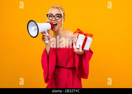 attractive blonde girl in a red dress shouts the news in a loudspeaker holding a gift box with a red ribbon on a yellow background Stock Photo