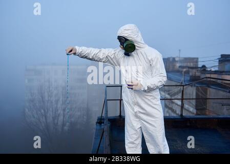 Scientist wearing protective outfit is conducting the study using blue reagent in glass flask on top of high building, contaminated area Stock Photo