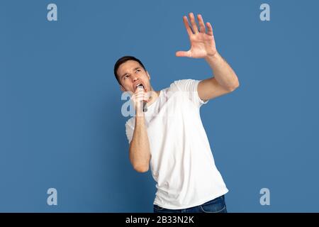 Singing like celebrities, stars. Caucasian young man's portrait on blue studio background. Beautiful male model in casual style, pastel colors. Concept of human emotions, facial expression, sales, ad. Stock Photo