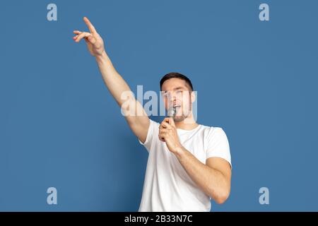 Singing like celebrities, stars. Caucasian young man's portrait on blue studio background. Beautiful male model in casual style, pastel colors. Concept of human emotions, facial expression, sales, ad. Stock Photo