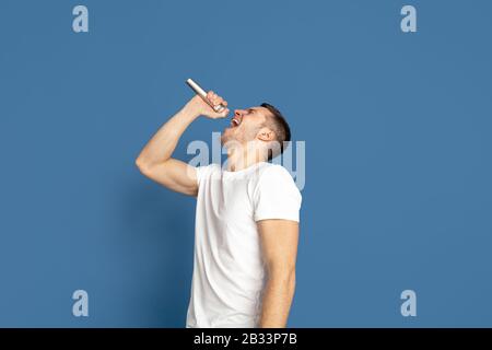 Singing like celebrities, stars. Caucasian young man's portrait on blue studio background. Beautiful male model in casual style, pastel colors. Concept of human emotions, facial expression, sales, ad. Stock Photo