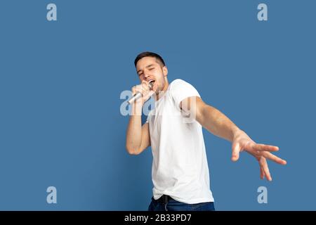 Singing like celebrities, stars. Caucasian young man's portrait on blue studio background. Beautiful male model in casual style, pastel colors. Concept of human emotions, facial expression, sales, ad. Stock Photo