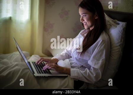 Asian woman using a notebook to work on night at the bed.She smiles and enjoys working at home. Stock Photo
