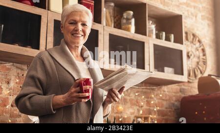 Cheerful Aged Woman Standing In Kitchen With Morning Coffee And Newspaper Stock Photo