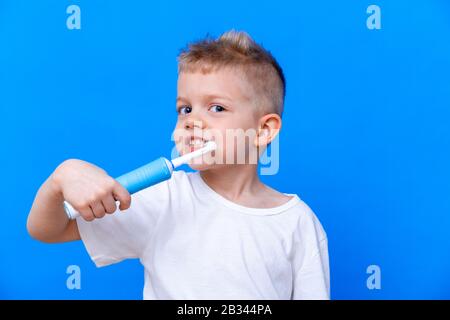 Happy child kid boy brushing teeth with electric toothbrush on blue background. Health care, dental hygiene. Mockup, copy space.  Stock Photo