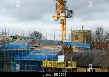 Construction site (roofers working, tiling roof, yellow crane, operator cab & mast, scaffolding) & church tower - York, North Yorkshire, England, UK. Stock Photo