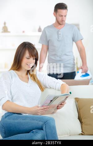 man doing some ironing while girlfriend is reading Stock Photo