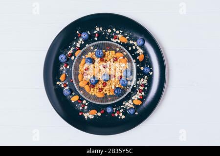 Healthy and delicious sweet breakfast of oatmeal granola with almonds and blueberries in glass bowl on black plate, top view on white table. Stock Photo