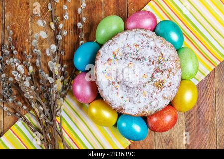 Easter decorations, background. Homemade Easter cake with colored eggs around it and willow branches on striped kitchen towel on wooden table. Top vie Stock Photo