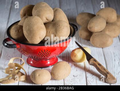 A potatoes in a drainer on a white wooden board on which there are other potatoes with one peeled potato and a knife Stock Photo