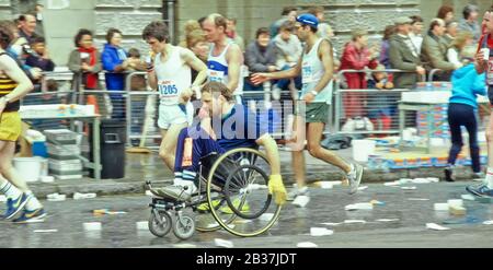 Disabled male athlete taking part in Mars sponsored London marathon wheelchair race passing able bodied athletes at water station Embankment road UK Stock Photo