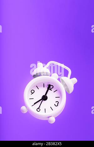 White retro style alarm clock in levitation isolated on purple background. Hands on the clock 7: 00, 19: 00. Minimalistic creative modern still life p Stock Photo