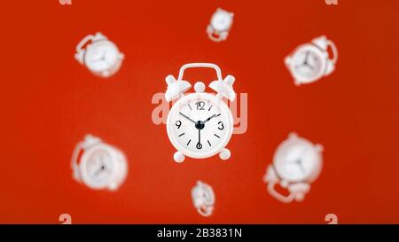 A white alarm clock in levitation in the center, isolated on a red background with an environment of hovering alarm clocks in the background. Creative Stock Photo