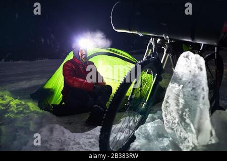 Bearded man with headlamp in the camp is sitting near bicycle and green tent at frozen lake in the mountains. Stock Photo