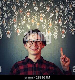 Joyful guy pointing index finger up, showing multiple light bulbs as creative ideas isolated on grey wall background. Positive thinking teenager, inge Stock Photo