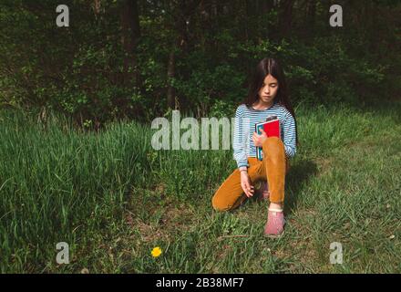 A thoughtful girl sits quietly with a book in tall grass in Springtime Stock Photo