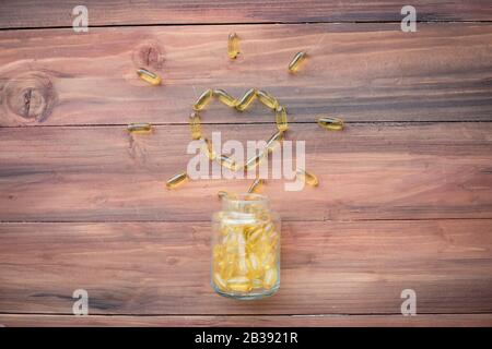 Pills in shape of heart dropped from bottle on wood background. Gold fish oil capsules. Concept of medicine in medical. Stock Photo