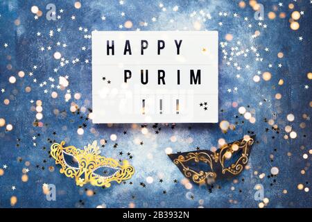 Flat lay of Purim Carnival celebration concept. Happy Purim written in light box, carnival mask and golden confetti on blue background. Top view Stock Photo