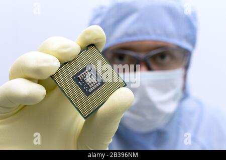 An engineer working in a clear room wearing a special uniform and protective glasses holds processor cpu in hand in glove Stock Photo