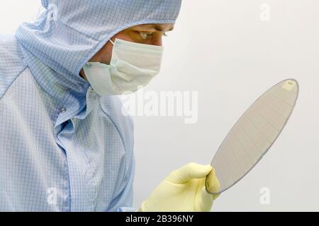 An engineer working in a clear room wearing a special uniform holds silicon wafer with microchips in hand in glove Stock Photo