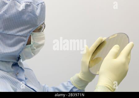 An engineer working in a clear room wearing a special uniform and protective glasses holds silicon wafer with microchips in hand in glove Stock Photo