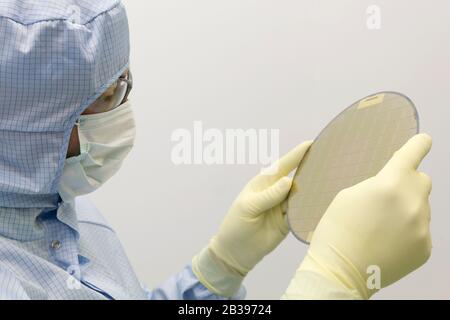 An engineer working in a clear room wearing a special uniform and protective glasses holds silicon wafer with microchips in hand in glove Stock Photo